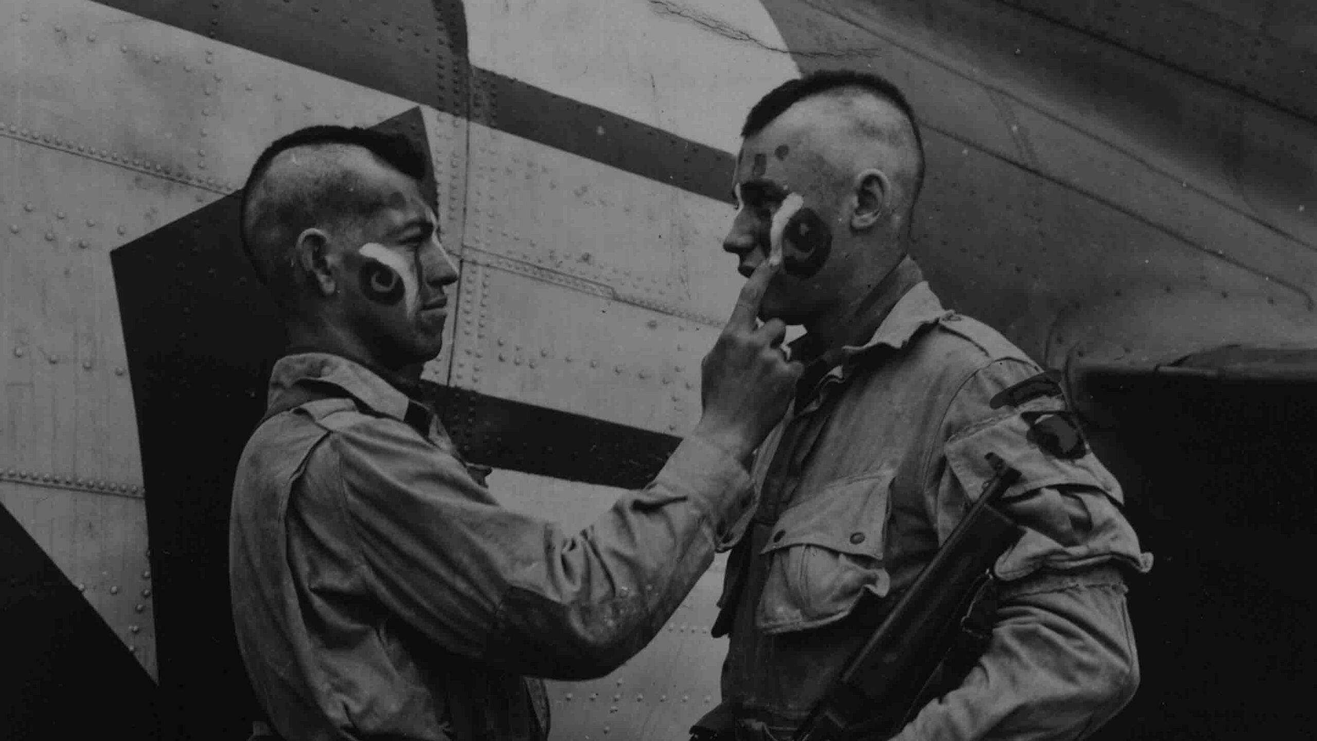 Private Clarence C. Ware applies final touches to the face paint of Private Charles R. Plaudo, styled after Native American patterns, in England, June 1944.